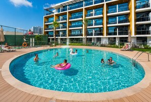 A swimming pool surrounded by a building with multiple balconies, a fenced area, and lounge chairs. - HAVN - Studio Apartment infront of CircleMall, JVC (Dubai)