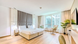 Moderni hotellihuone, jossa on suuri sänky, oleskelualue tuolilla ja pienellä pöydällä sekä suuri ikkuna, josta avautuu näkymä kaupunkiin.
