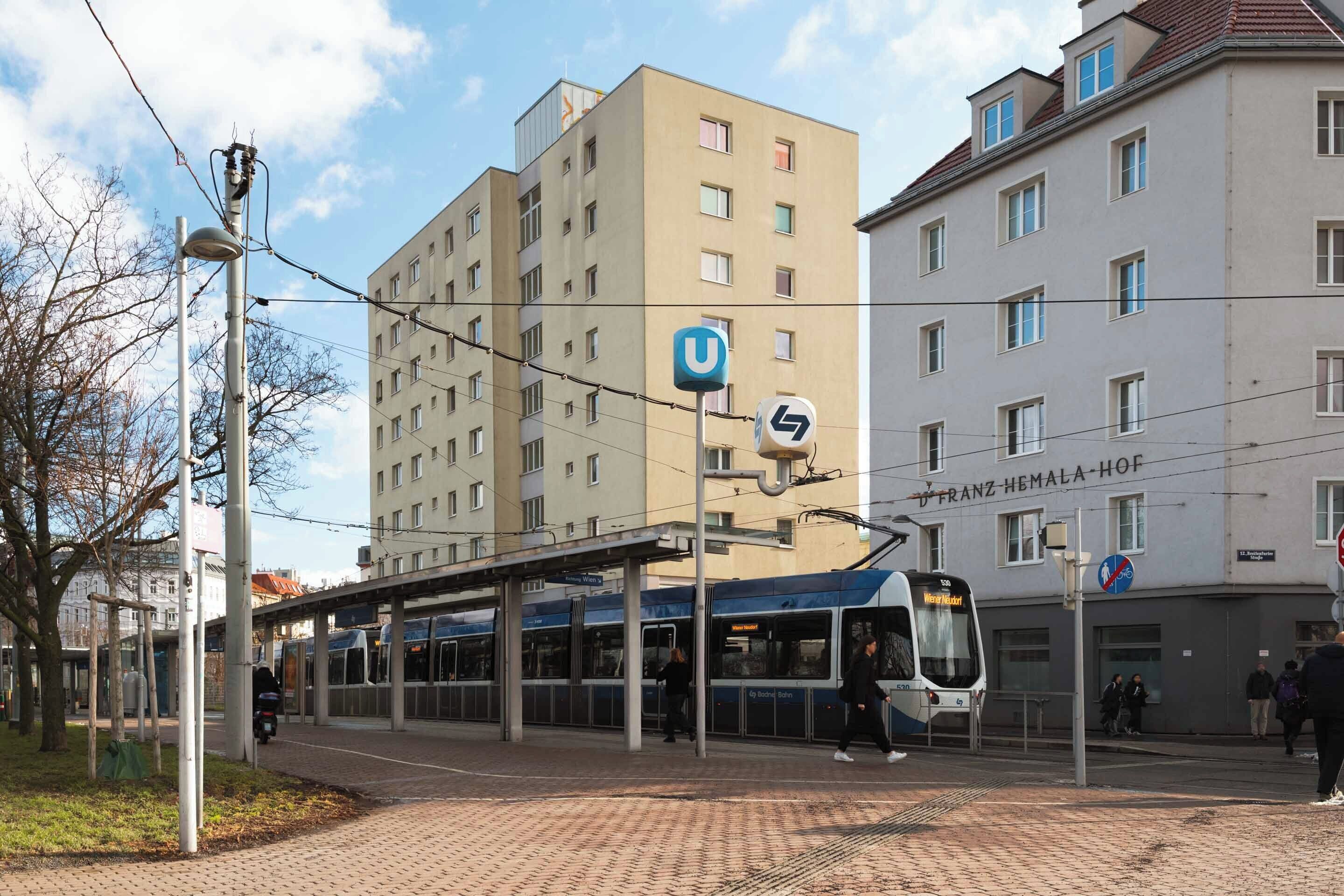 Una estación de tranvía con un tranvía, un cartel en forma de U y un edificio con la señalización 'FRANZ HEMALA-HOF'.