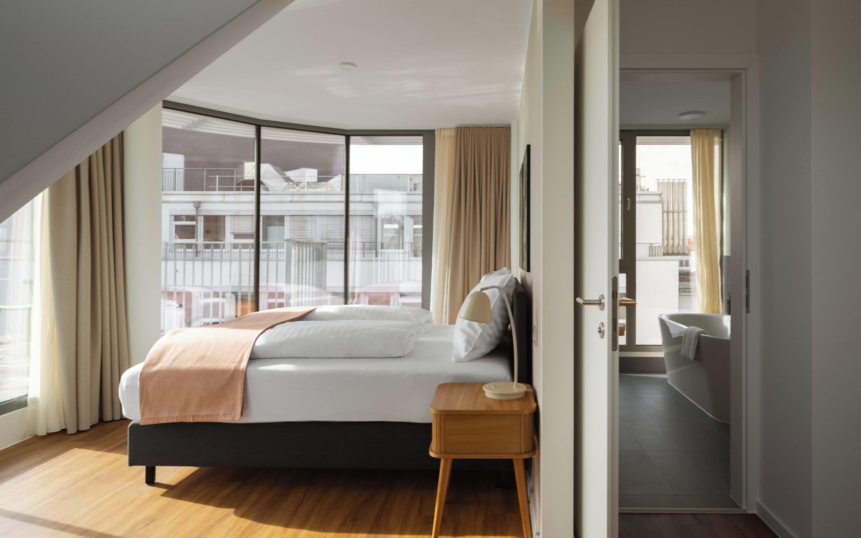 Ett modernt hotellrum med en stor säng, ett träbord som sängbord och utsikt över staden genom stora fönster som sträcker sig från golv till tak.