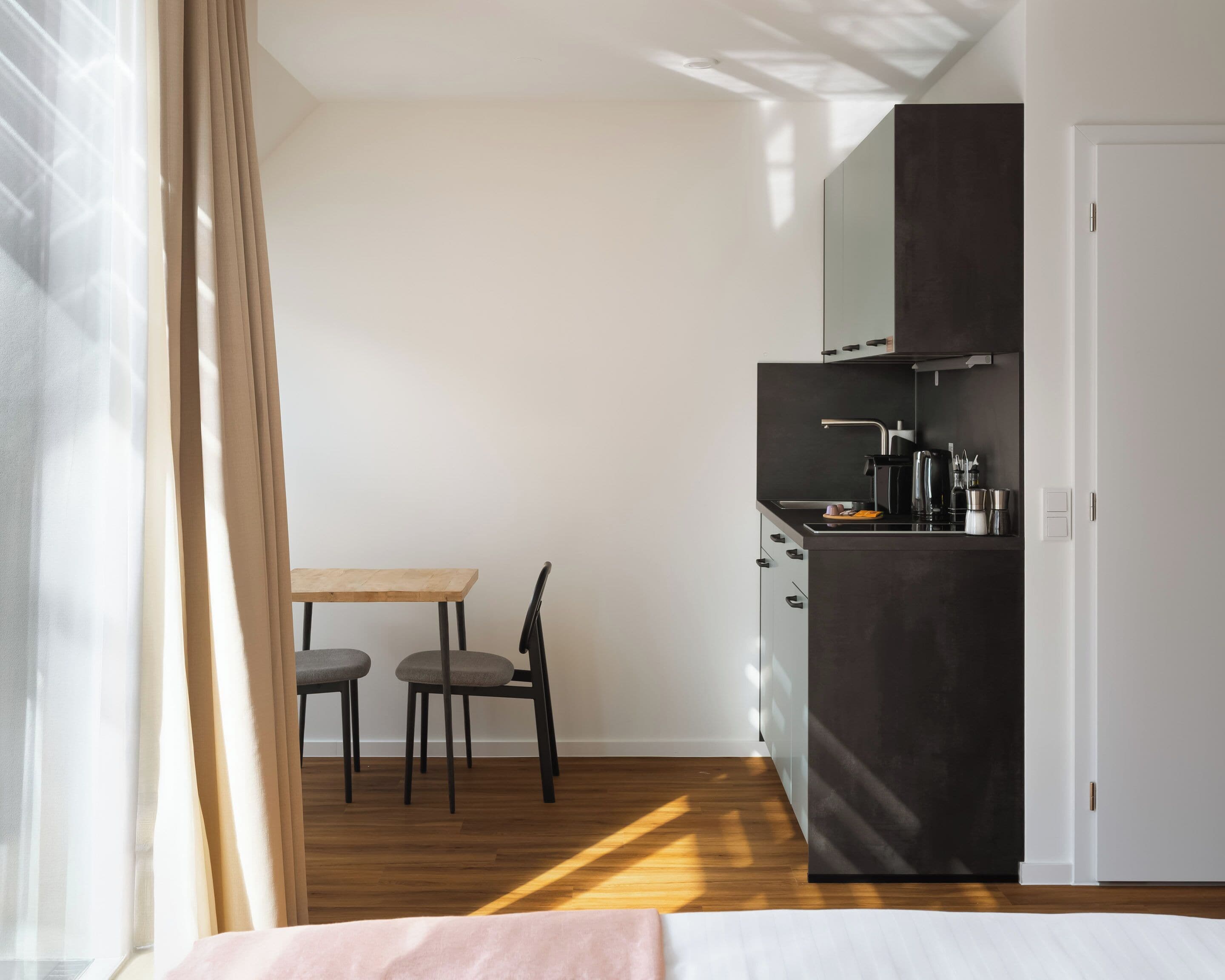 A compact room with a bed, a small table with two chairs, a kitchenette, and a window with curtains.