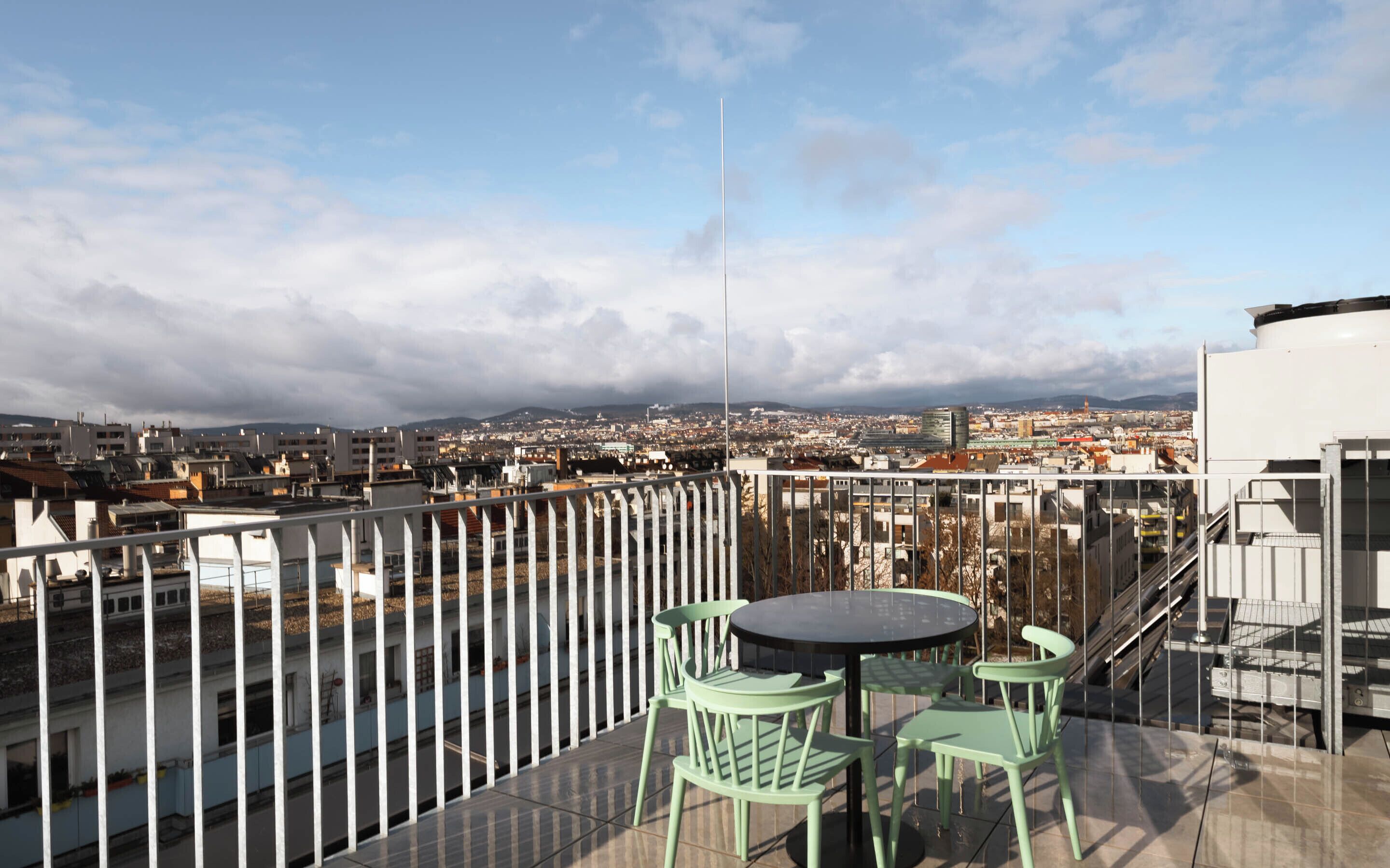 Eine Dachterrasse mit Tisch und Stühlen mit Blick auf eine Stadtlandschaft, einschließlich Gebäude und einem bewölkten Himmel.