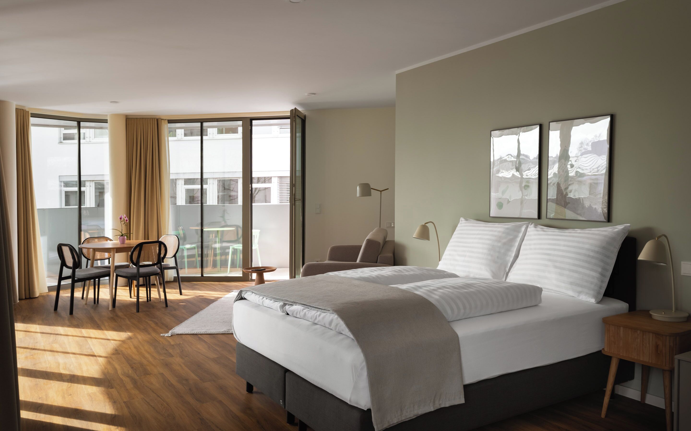 A modern hotel room with a large bed, a desk with chairs, a balcony with a table and chairs, and artwork on the walls.