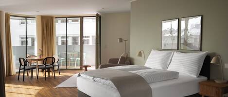 A modern hotel room with a large bed, a desk with chairs, a balcony with a table and chairs, and artwork on the walls.