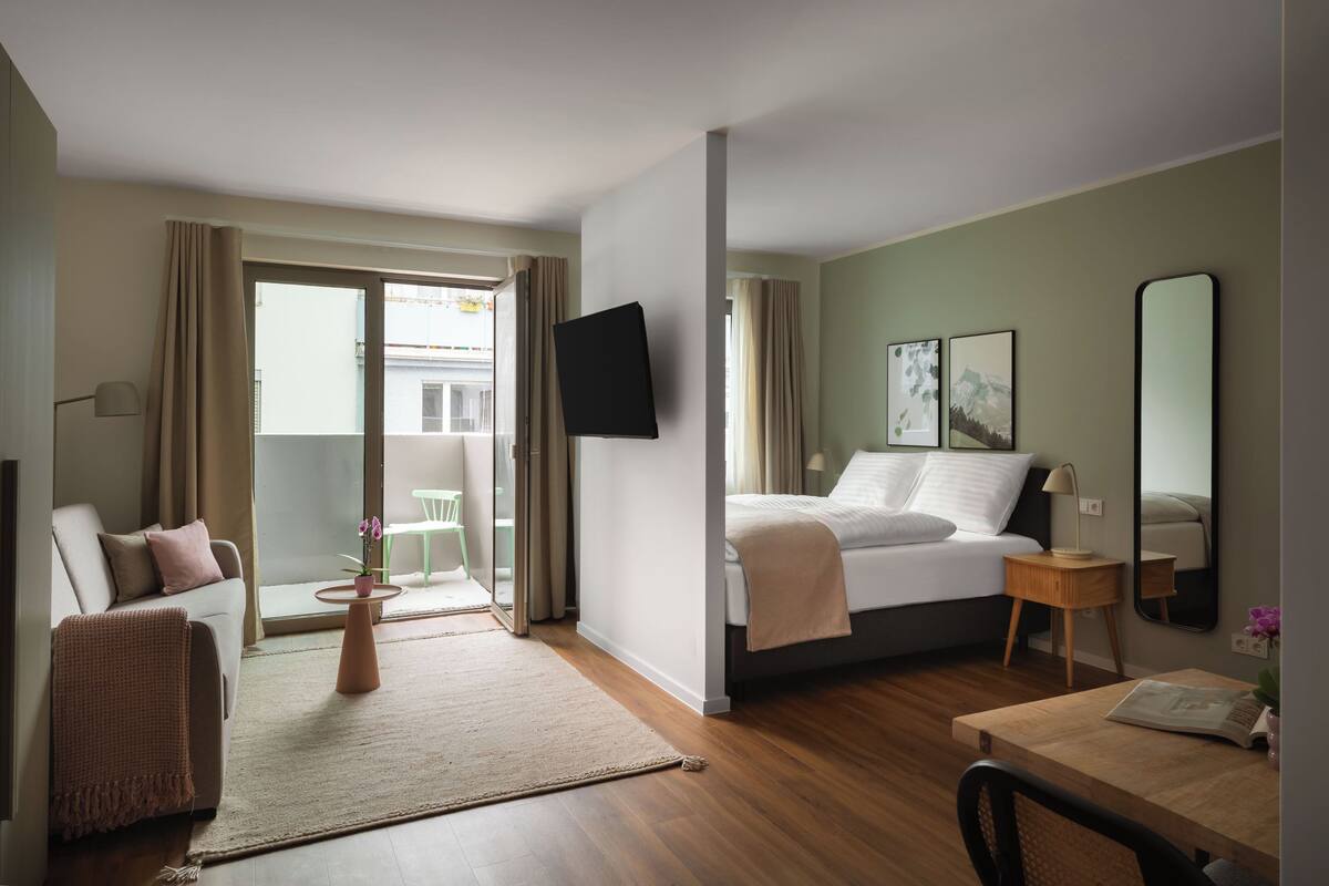 Ein Hotelzimmer mit Bett, Schreibtisch, Stuhl, Fernseher und einem Balkon mit Blick auf Gebäude.