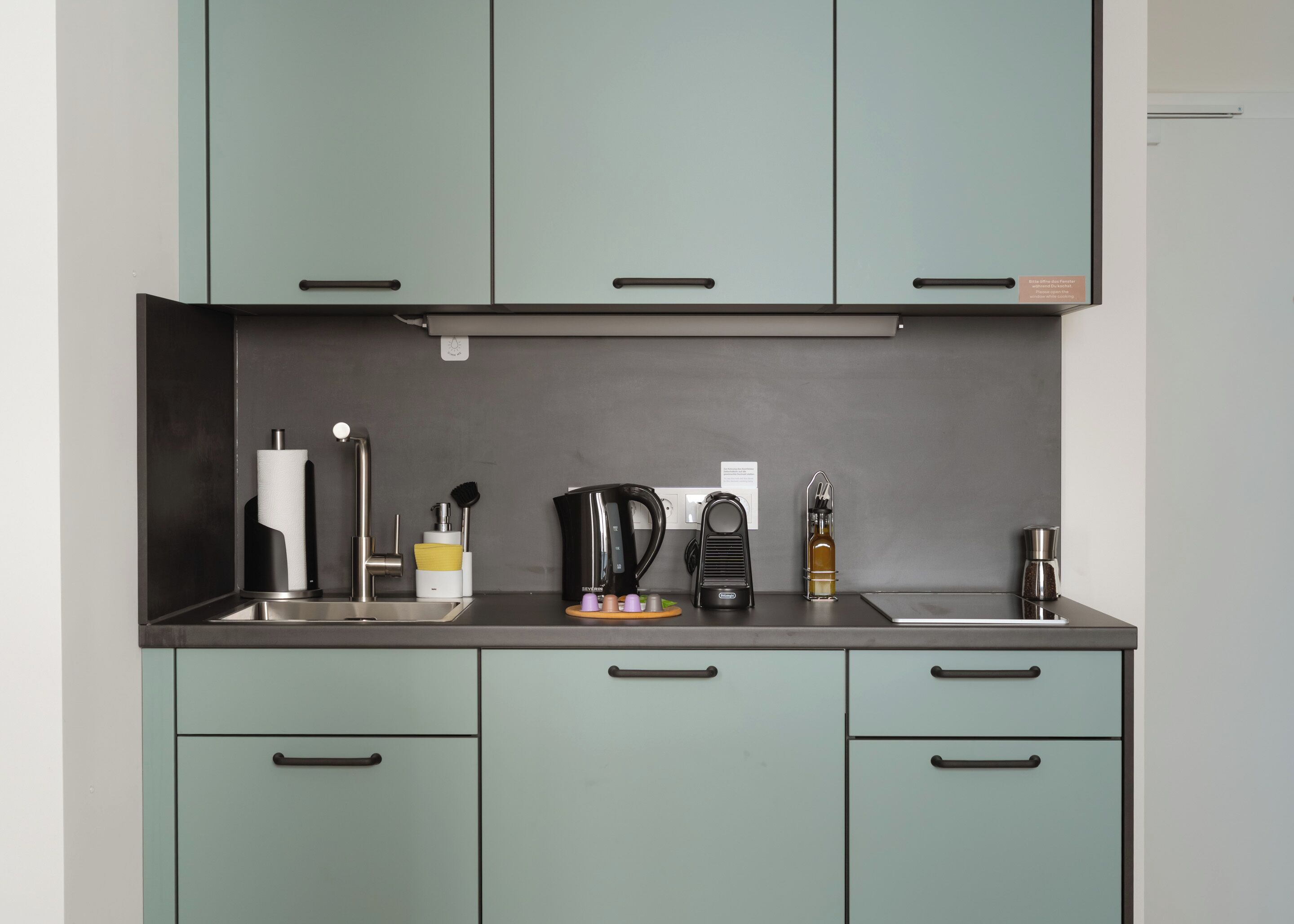 A modern kitchen with teal cabinets, a stainless steel countertop, and various appliances including a coffee maker and a kettle.