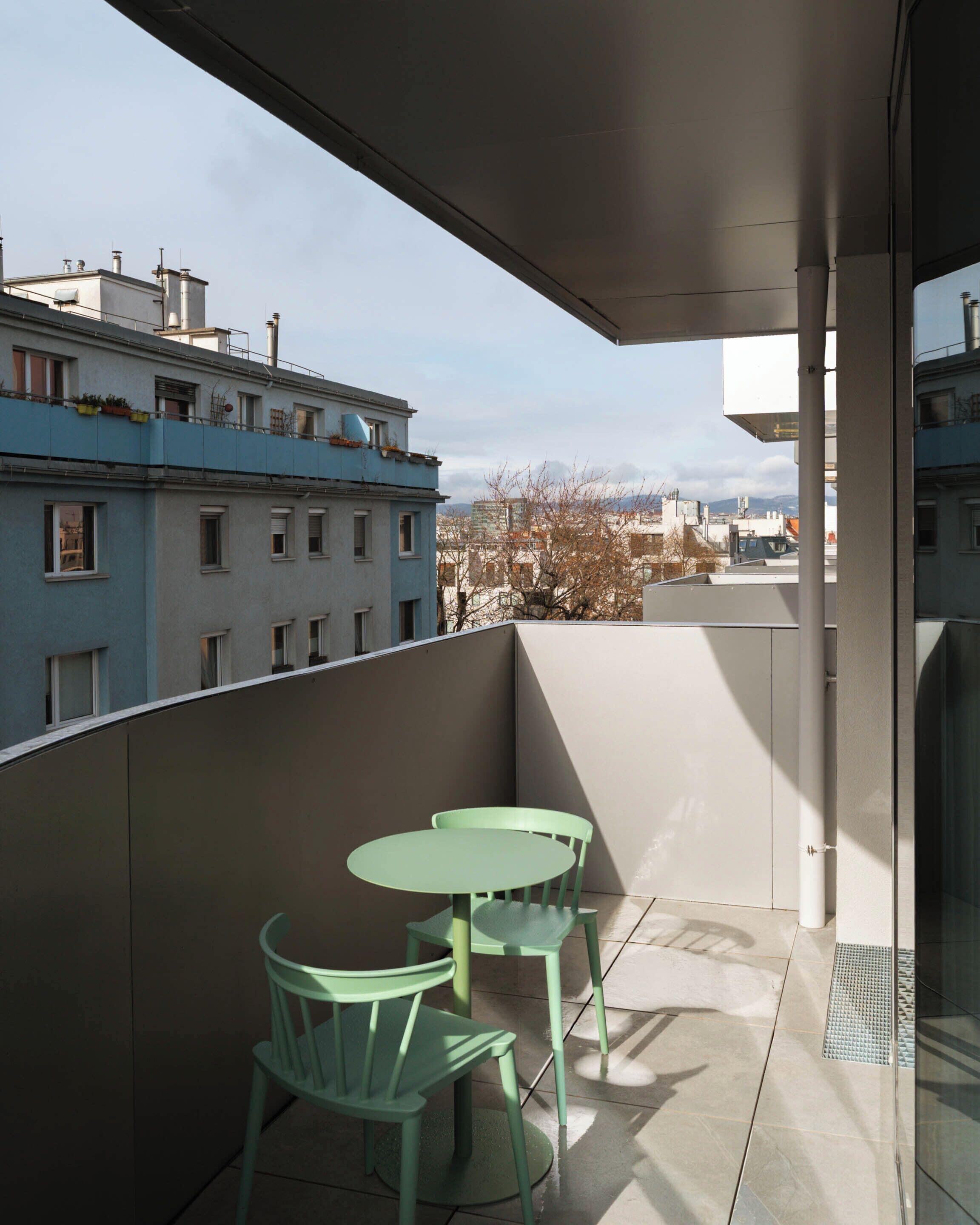 A balcony with a table and two chairs, overlooking a cityscape with buildings and a clear sky.
