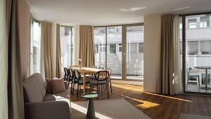 Una sala de estar moderna con sofá, mesa de comedor y sillas, ventanas amplias con cortinas, y un balcón con mobiliario exterior.