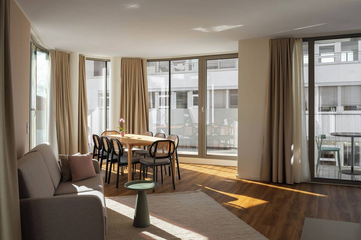 Ein modernes Wohnzimmer mit einer Couch, einem Esstisch, Stühlen, großen Fenstern mit Vorhängen und einem Balkon mit Gartenmöbeln.