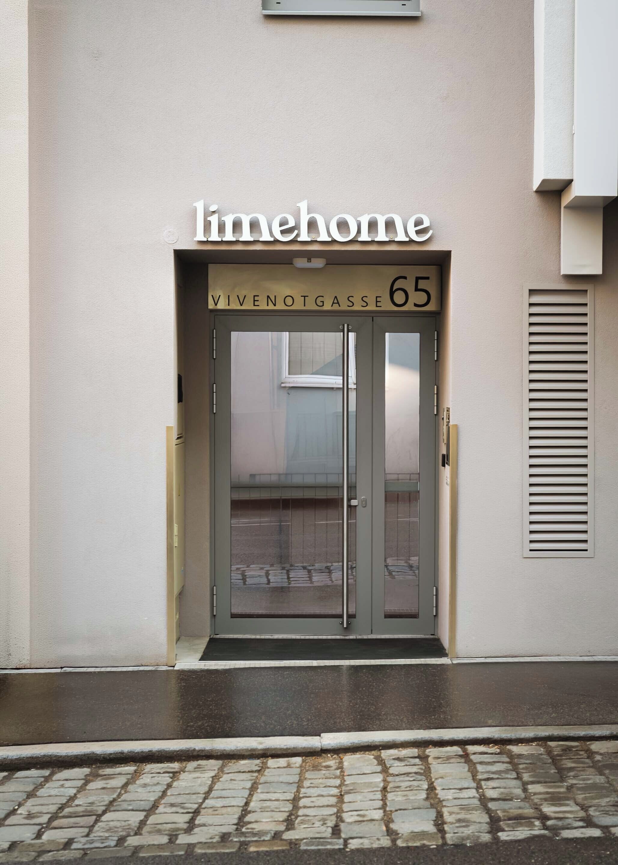 Una entrada de edificio con puerta de cristal, un letrero que dice "limehome" arriba y la dirección "WIVENOTGASSE 65".