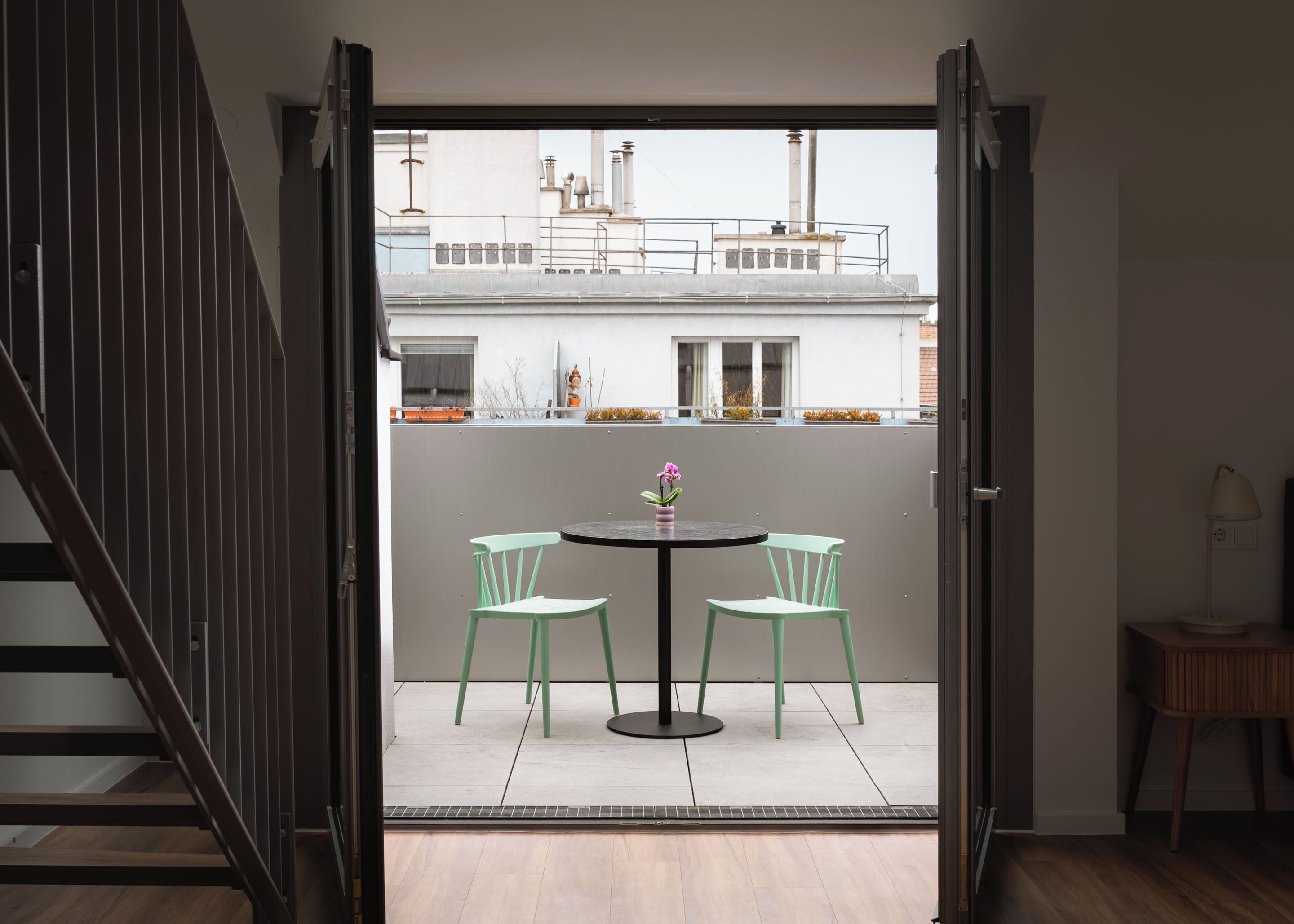 Een geopende deur die leidt naar een balkon met een tafel, twee stoelen, een potplant en een uitzicht op gebouwen op de achtergrond.