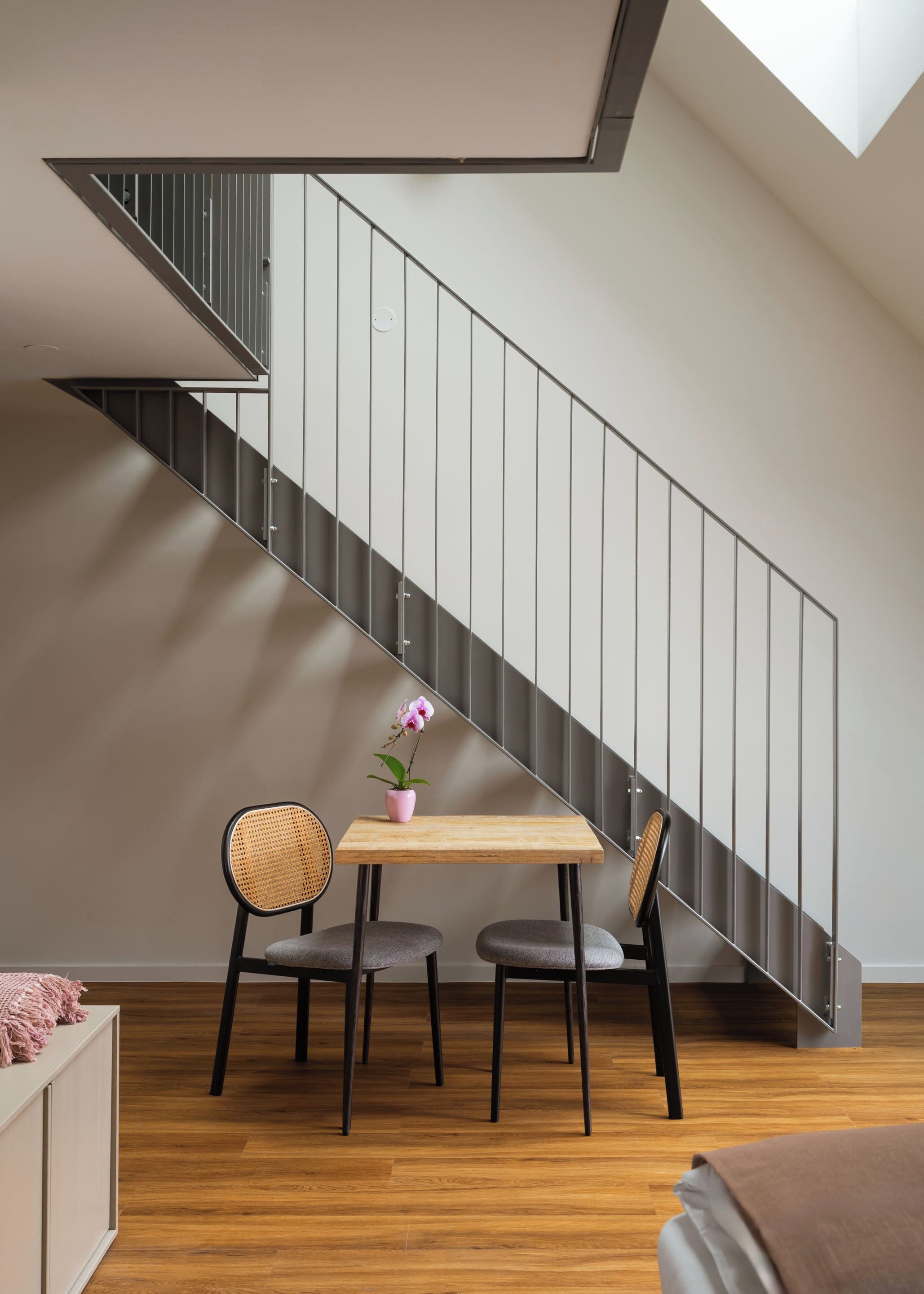 A modern interior with a wooden table, two chairs, and a potted orchid. A staircase with metal railings is visible in the background.