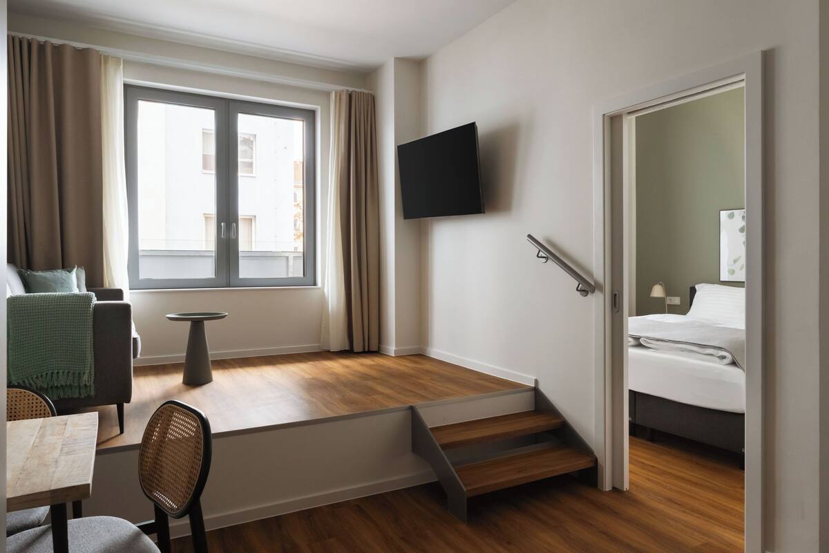 Ein modernes Hotelzimmer mit Holzboden, einem Wand-Fernseher, einem Bett mit weißer Bettwäsche, einem kleinen Tisch mit Stuhl und einem Fenster mit Vorhängen.