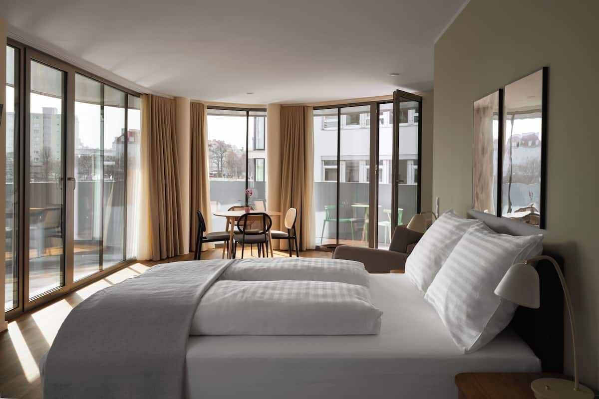 Ein Hotelzimmer mit einem großen Bett, einem Esstisch mit Stühlen und großen Fenstern mit Blick auf die Stadt.