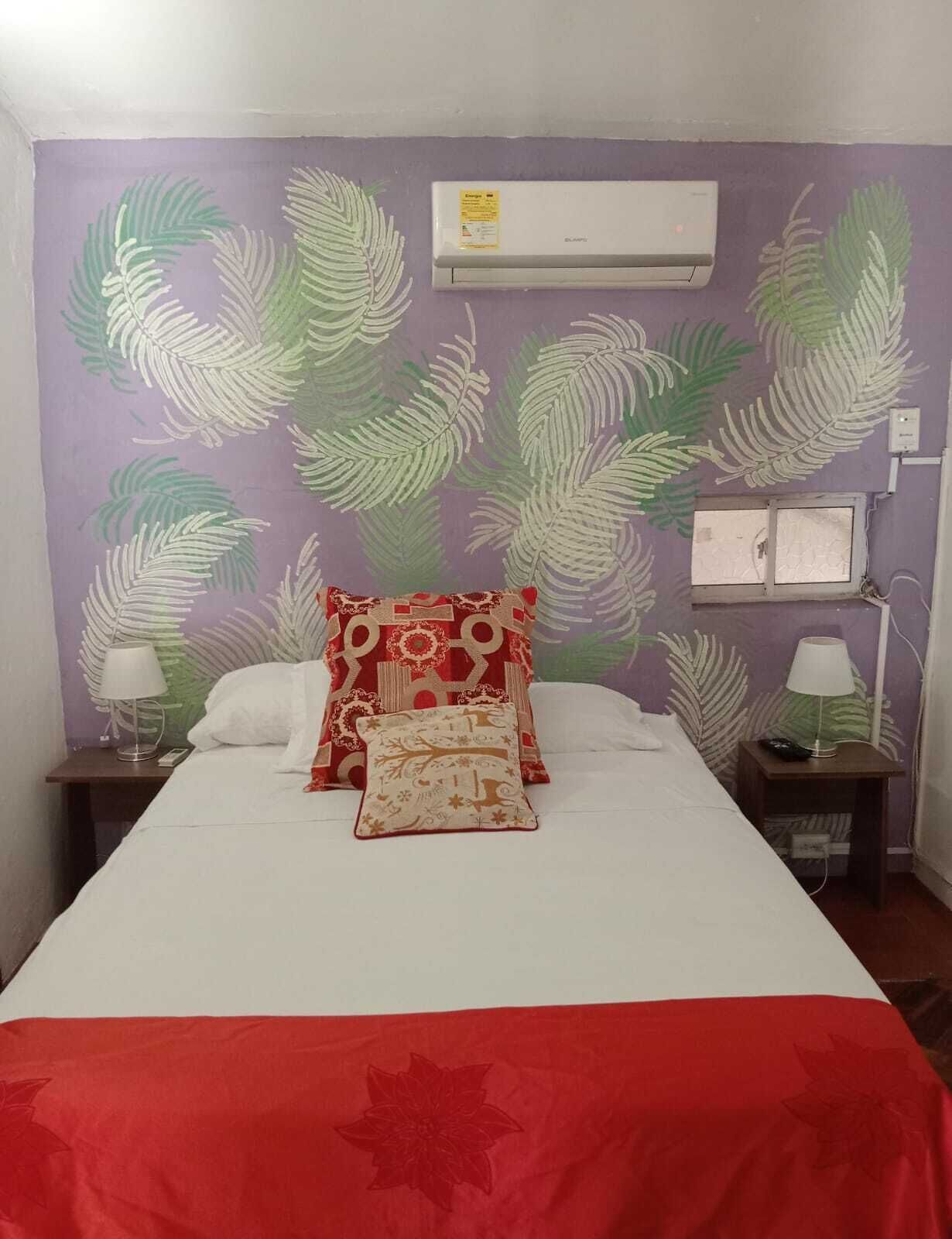 A hotel room with a bed, a red and white patterned pillow, a white pillow, a red and white blanket, an air conditioner, a nightstand with a lamp, and a wall with a leaf pattern.
