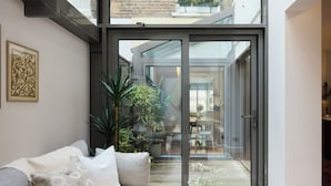 A modern living room with a sliding glass door leading to a balcony, a sofa with cushions, a coffee table, and a potted plant. - The Kensington End House (London)
