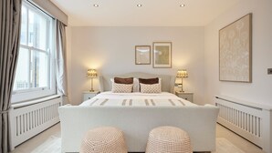A bedroom with a large bed, two bedside lamps, a window with curtains, and a wall with two framed pictures. - The Kensington End House (London)
