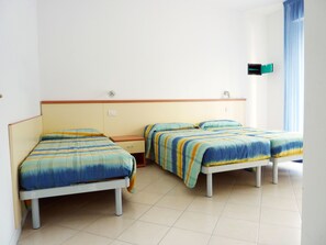A room with three single beds, each with a striped bedspread. There is a small wooden nightstand between the beds, and a window with blue curtains on the right side. - Cozy beachside apartment (Bibione Pineda)
