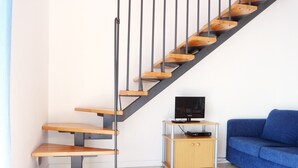 A staircase with wooden steps and metal railings, a small wooden cabinet with a television, and a blue sofa in a room with tiled flooring.