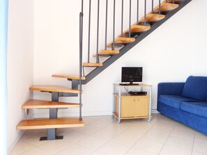 Eine Treppe mit Holzboden und Metallgeländern, eine kleine Holzschublade mit einem Fernseher und ein blaues Sofa in einem Raum mit Fliesenboden.