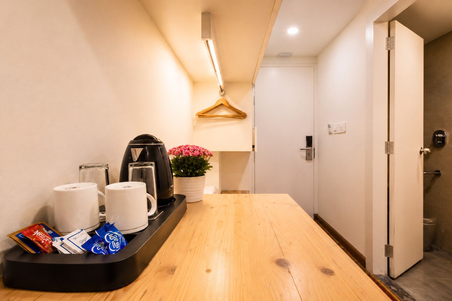 A hotel room with a wooden table, a kettle, a coffee maker, a tray with snacks, a potted plant, and a door leading to a bathroom.
