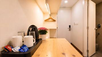 A hotel room with a wooden table, a kettle, a coffee maker, a tray with snacks, a potted plant, and a door leading to a bathroom.