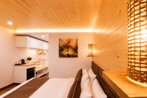 Een moderne hotelkamer met een houten plafond, een bed met witte lakens, een schilderij aan de muur en een klein keukenhoekje.