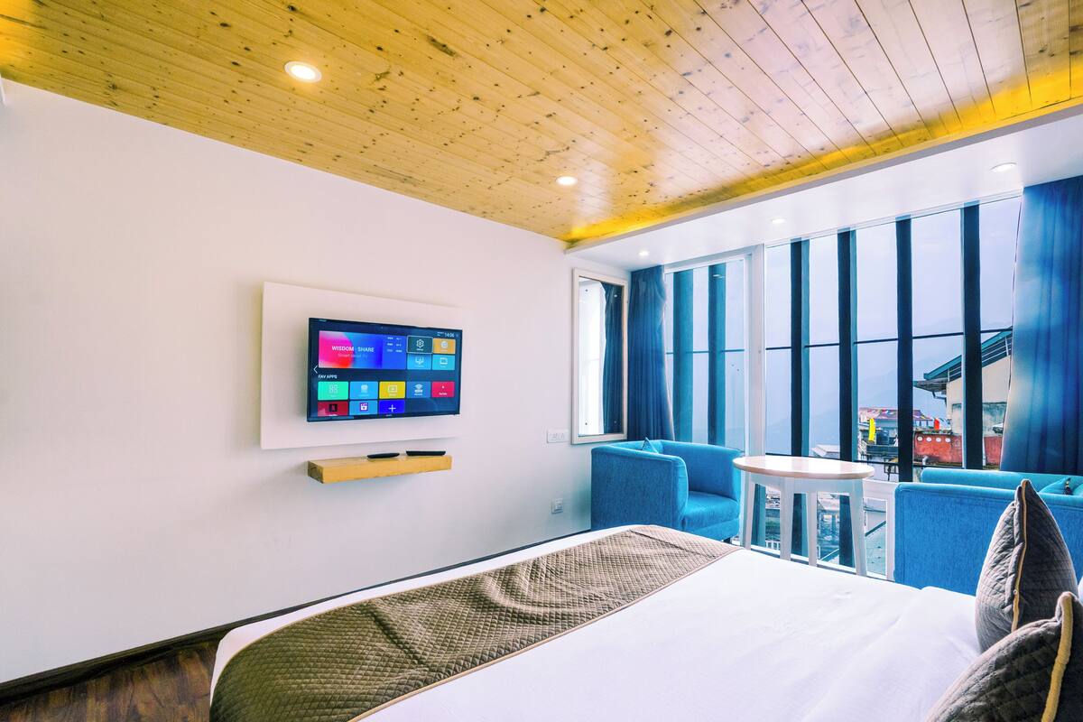 Habitación de hotel moderna con techo de madera, un ventanal con vista a la ciudad, un televisor de pantalla plana y una zona de estar con sillas azules.