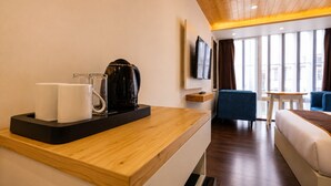 Habitación de hotel con escritorio de madera, cafetera, televisor montado en la pared, cama y zona de estar con una silla azul y una mesita.