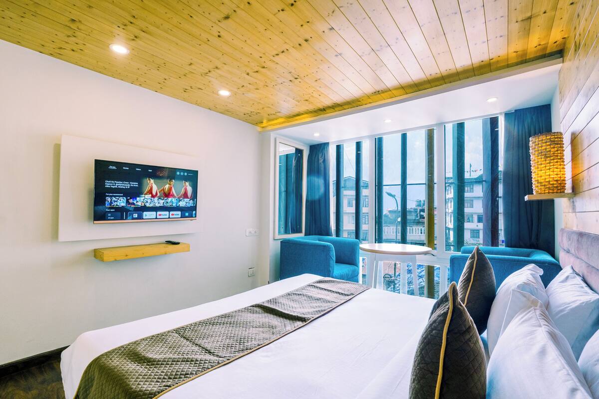 Una habitación de hotel moderna con una cama grande, un televisor montado en la pared, una ventana con cortinas, una mesita pequeña y una silla azul.