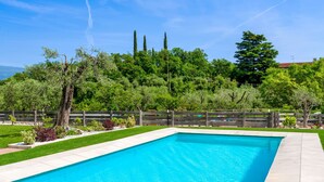 Una piscina rectangular con solárium empedrado, rodeada por una valla de madera y abundante vegetación.
