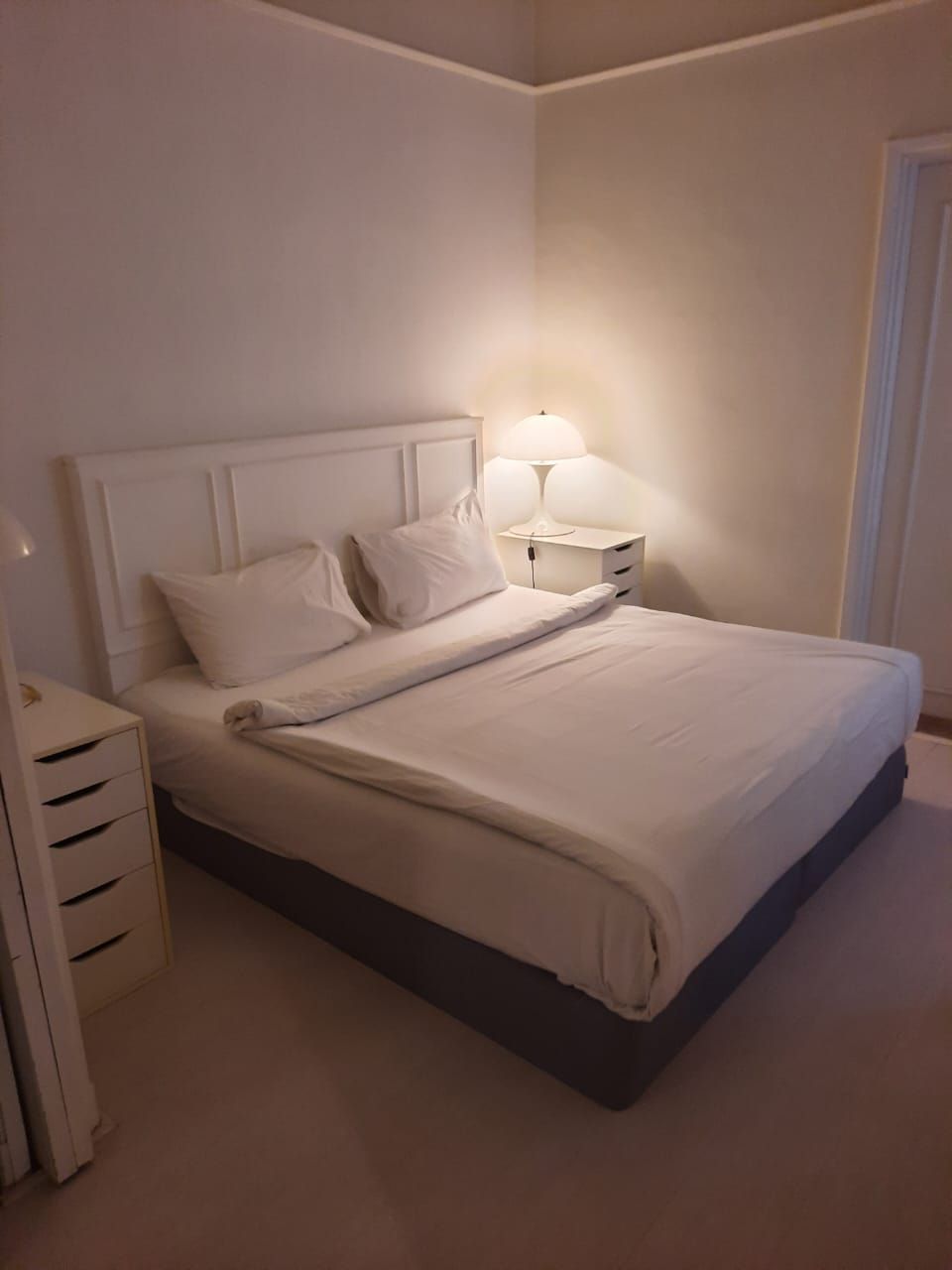 A neatly made bed in a hotel room with a white headboard, a bedside table with a lamp, and a small drawer unit.