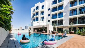 A modern building with a swimming pool, outdoor seating, and a clear sky. - HAVN - Studio Apartment in Pantheon Elysee, JVC (Dubai)