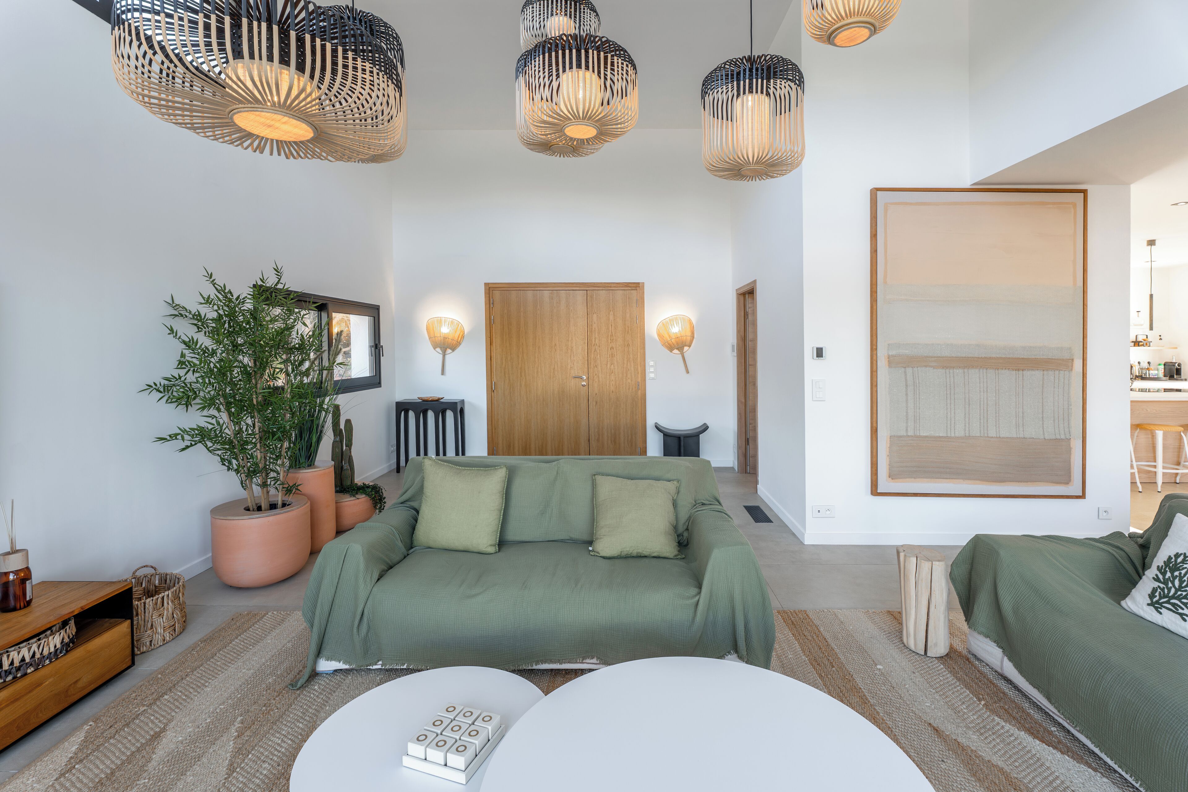 A modern living room with a green sofa, a white coffee table, and a large window. There are potted plants and a wooden door in the background.