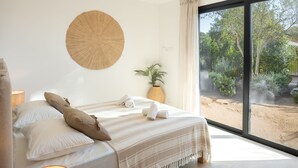 A bedroom with a large bed, a round wall hanging, a potted plant, and a view of a garden through a large window. - VILLA JUCA - SAINT CYPRIEN SOUTH CORSICA- 10 PERS- 5 MIN FROM THE BEACH (Zonza)