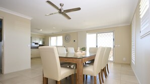 Ein Essbereich mit einem Tisch mit Glasplatte, sechs weißen Stühlen, einem Deckenventilator und einer Küche im Hintergrund.