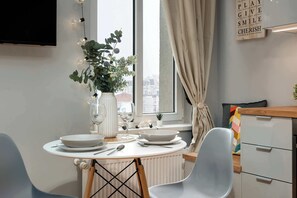 Uma área de refeições com uma mesa redonda posta para dois, um vaso com flores e uma janela com cortinas.
