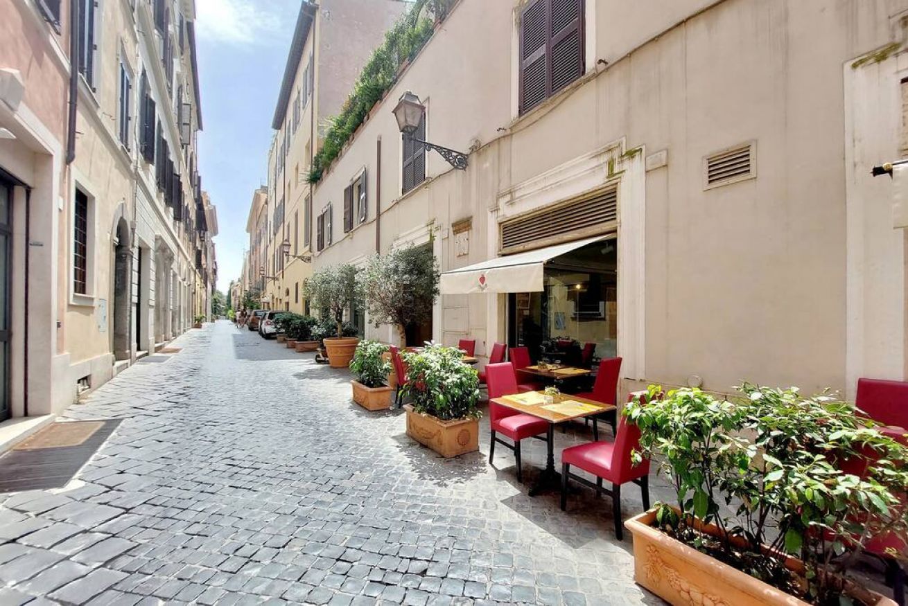 Une ruelle étroite pavée, bordée d’immeubles, avec des terrasses dotées de chaises et de tables rouges, et des plantes en pot.