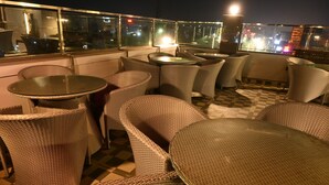 A rooftop terrace with wicker furniture and glass tables, overlooking a cityscape at night. - Hotel Aveda Paradise (Varanasi)