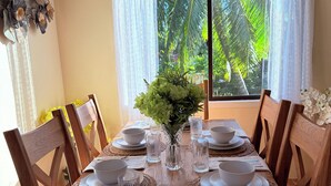 A dining table set for six with white plates, bowls, and glasses, a vase with greenery, and wooden chairs.