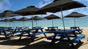 A beach with blue picnic tables and black umbrellas with Heineken logos.