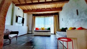 A room with a wooden ceiling, a bed with colorful pillows, a white sofa, a red stool, and a TV on the wall. - Sunflowers abode Your rustic home in the center of all Tuscan destinations! (Toscana)