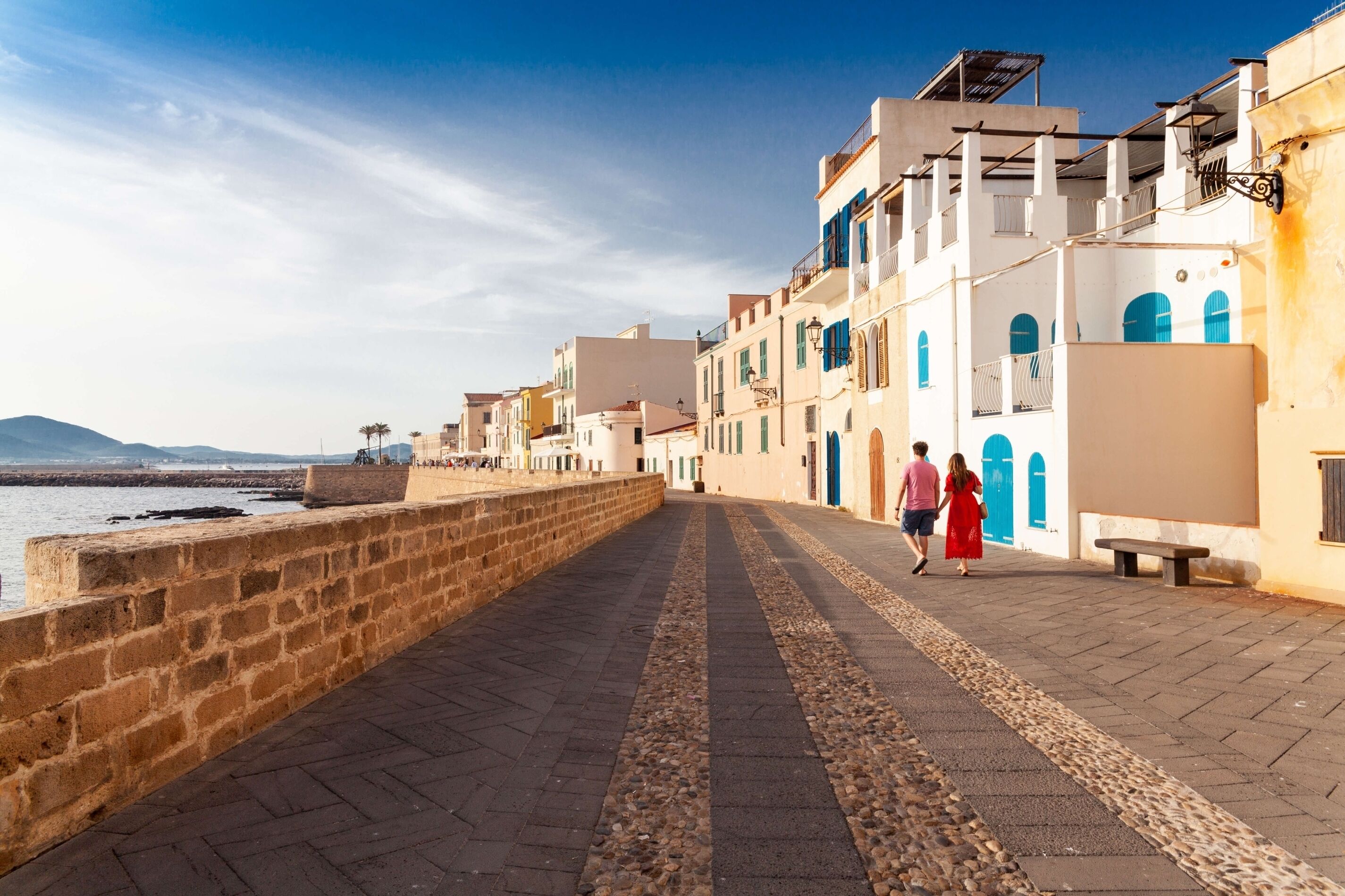 A coastal promenade with buildings, a stone wall, and a clear sky.