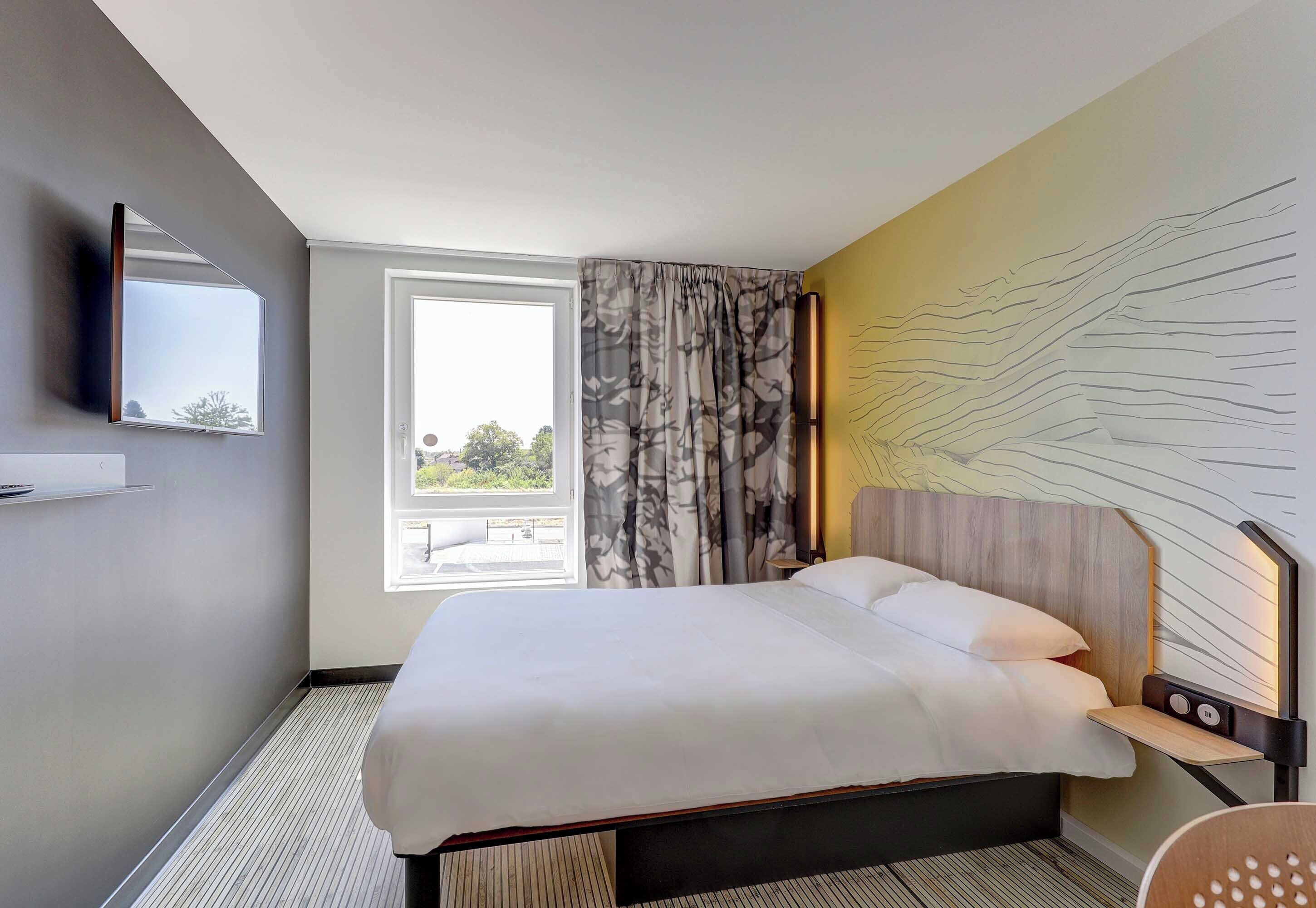Ein Hotelzimmer mit Bett, Wandfernseher, Fenster mit Vorhängen und einem Wandbild.