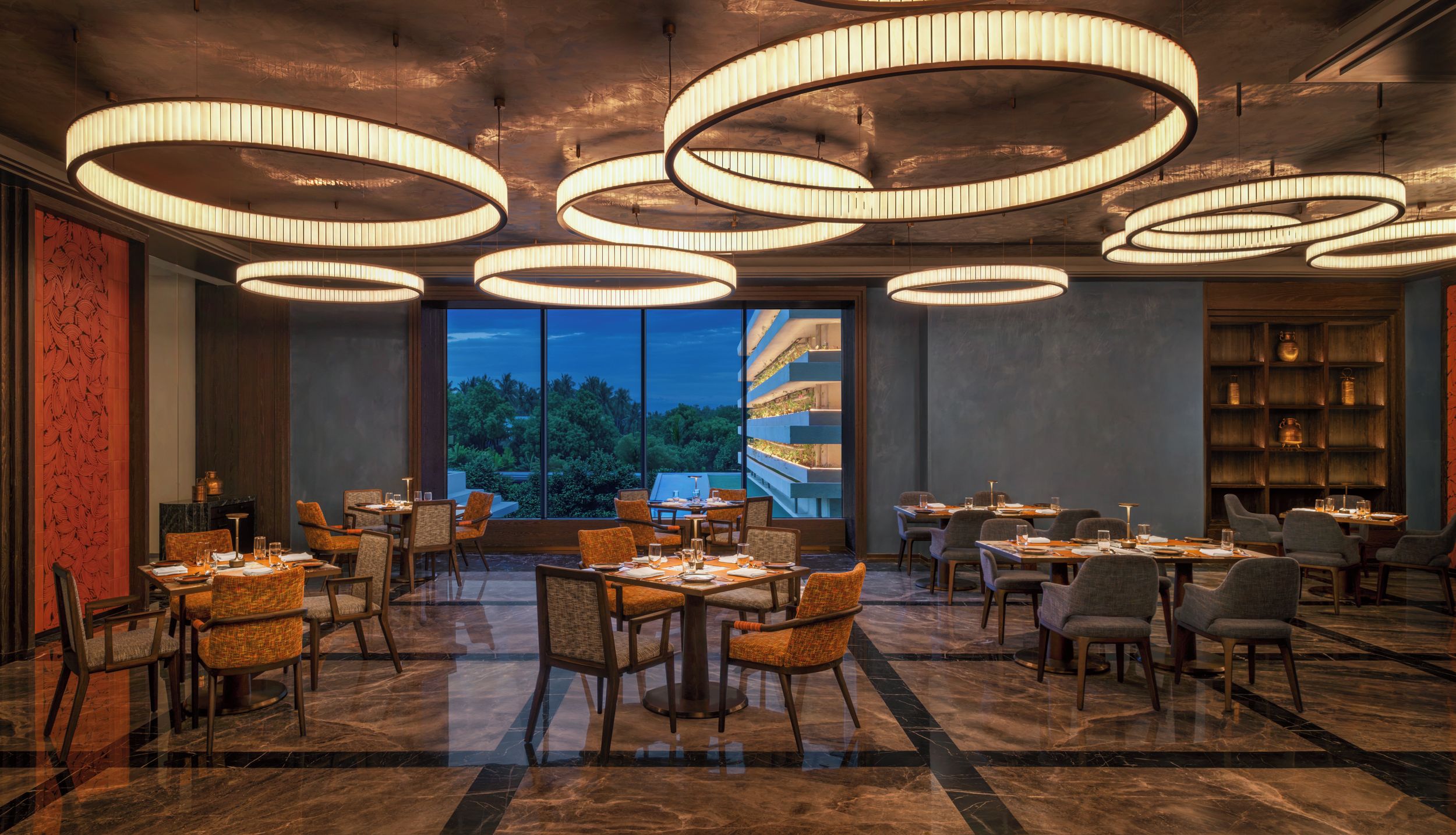 A spacious dining area with multiple tables and chairs, large windows offering a view of greenery, and modern circular ceiling lights.