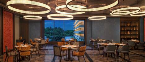 A spacious dining area with multiple tables and chairs, large windows offering a view of greenery, and modern circular ceiling lights.