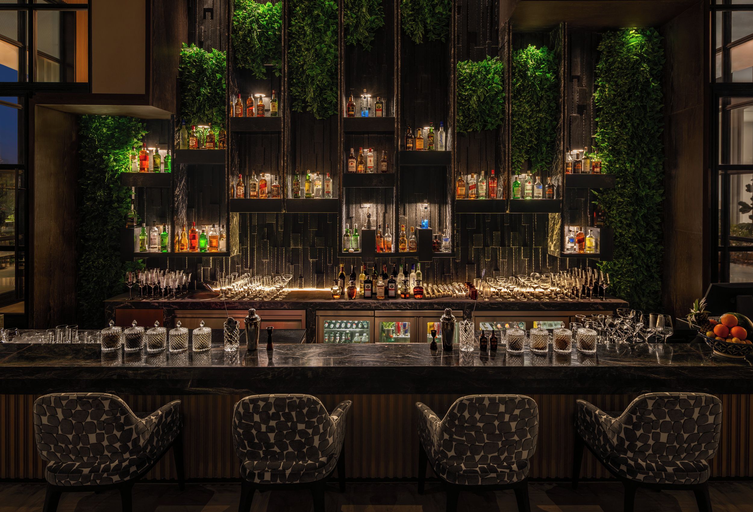A bar area with multiple shelves of liquor bottles, a bar counter with glasses, and three patterned chairs.