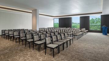 Une salle de conférence avec des rangées de chaises, un pupitre et de grandes fenêtres donnant sur l’extérieur.