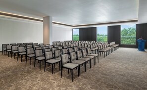 Un salón de conferencias con filas de sillas, un atril y grandes ventanales que ofrecen vistas al exterior.