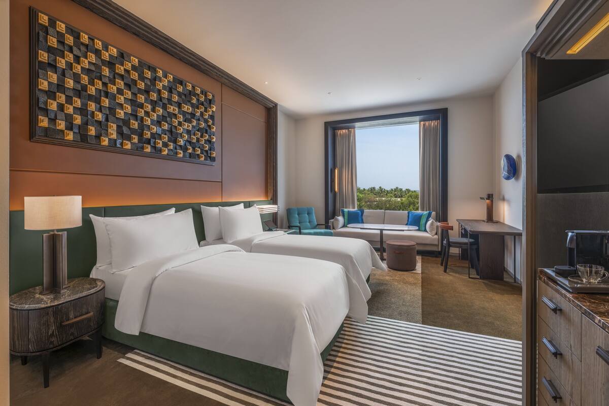 A hotel room with two beds, a dining area, a view of the outdoors, and a large decorative wall.